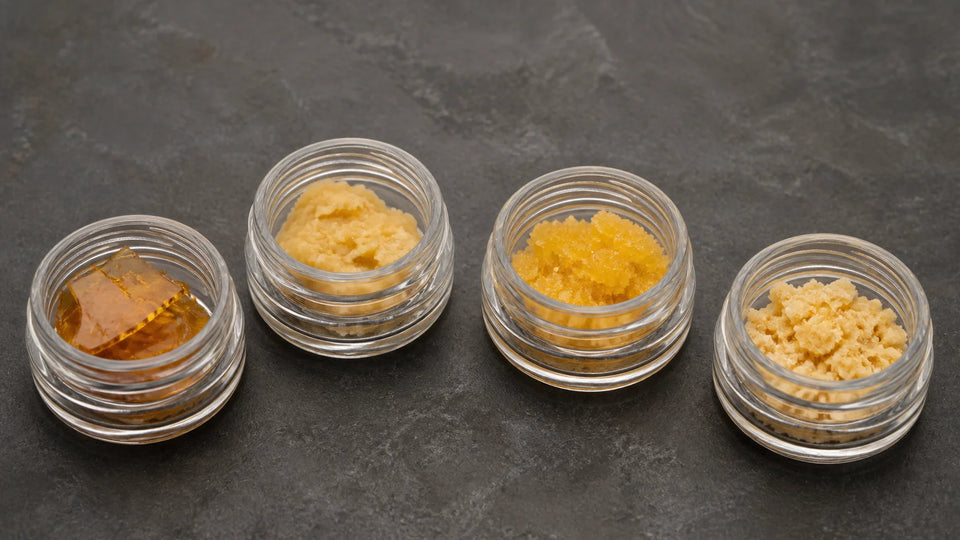 Four identical glass jars show shatter, budder, sugar, and crumble concentrate textures on one dark tabletop.